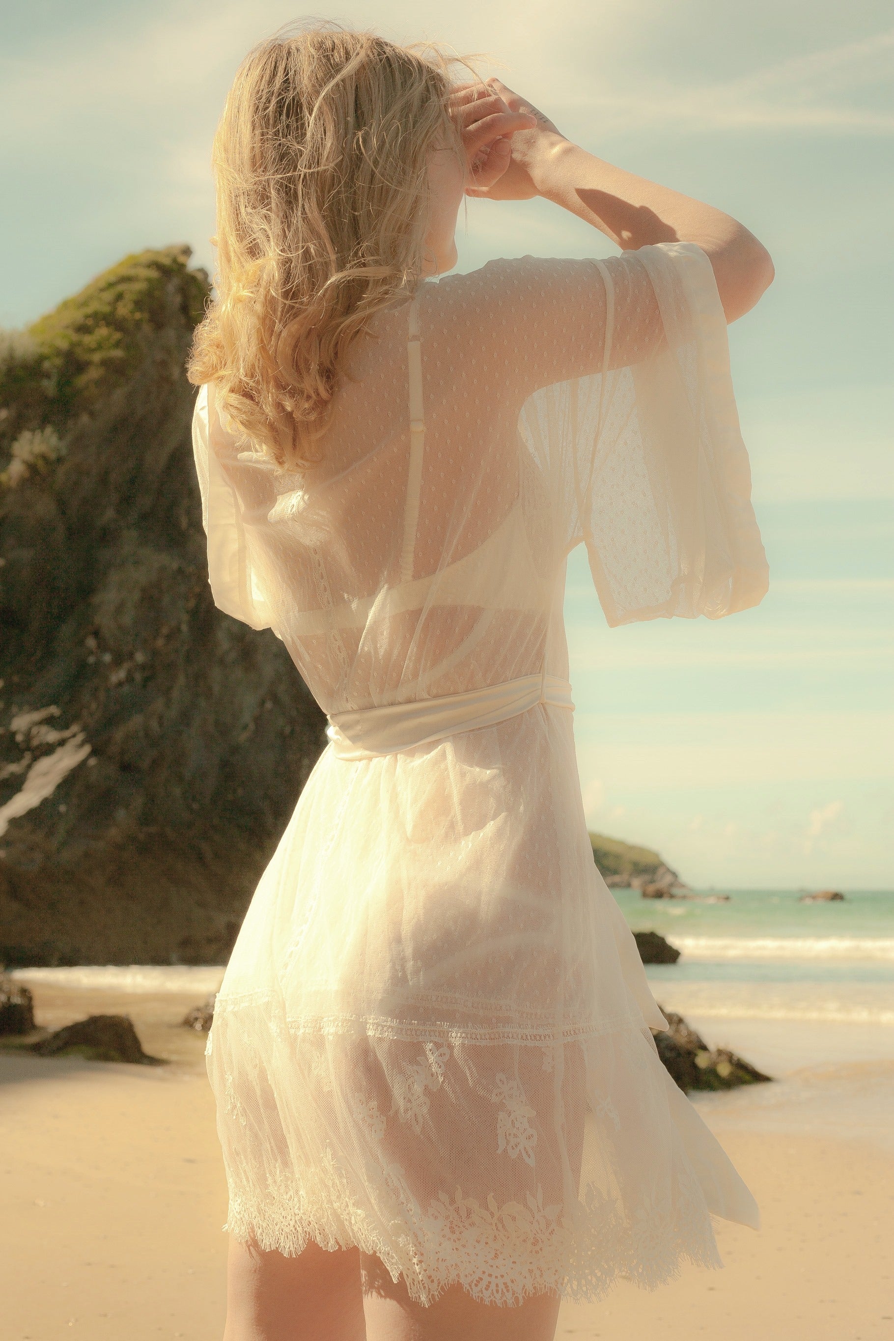 Woman in a white lace dress standing on a beach with ocean and rocks in the background
